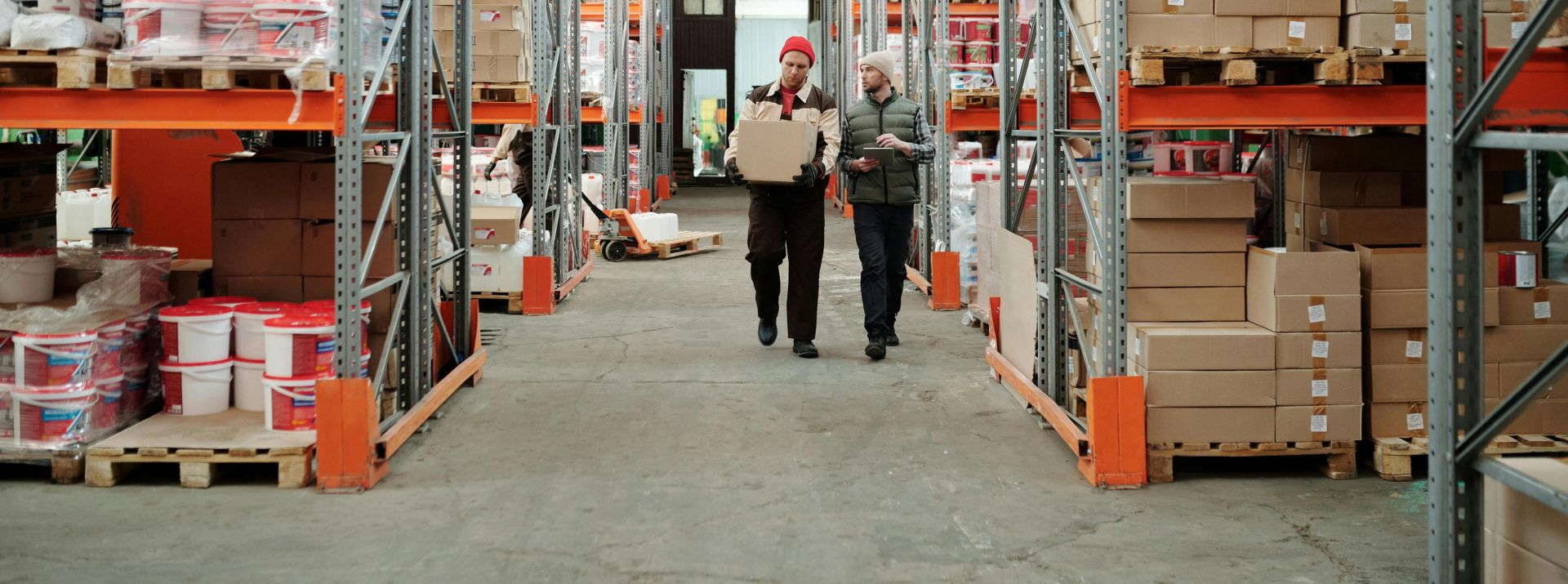 Two workers handle a package in a spacious warehouse surrounded by shelves stocked with boxes and products.